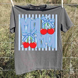 May include: Gray t-shirt with a graphic design featuring the word "MOM" in blue gingham print, adorned with red cherries, blue bows, and white flowers. The shirt is hanging on a clothesline.