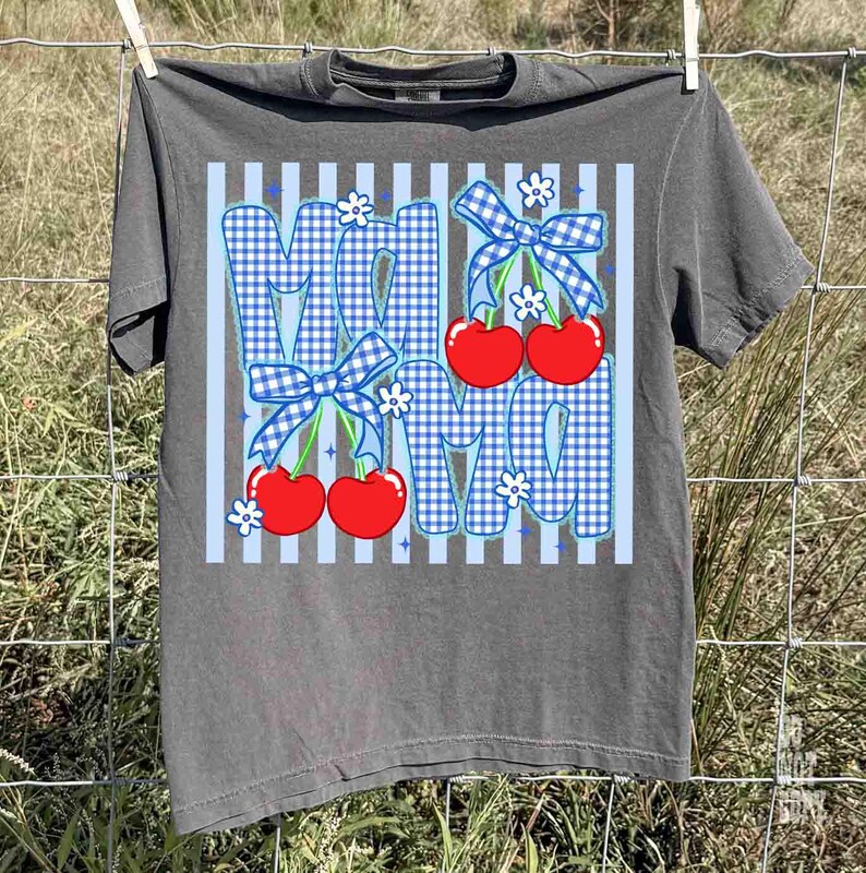 May include: Gray t-shirt with a graphic design featuring the word "MOM" in blue gingham print, adorned with red cherries, blue bows, and white flowers. The shirt is hanging on a clothesline.