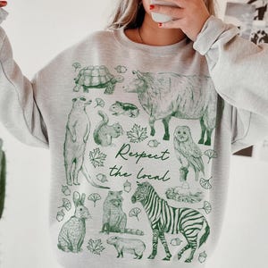 May include: A light gray sweatshirt featuring a green illustration of various animals, including a zebra, owl, and squirrel. The text "Respect the local" is also printed on the sweatshirt.