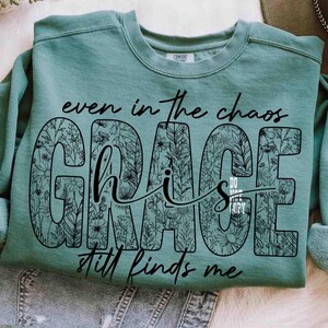 May include: A teal-colored sweatshirt with the words "even in the chaos GRACE his GRACE still finds me" in black script and floral design. The sweatshirt is made of a soft material and has a crew neck.
