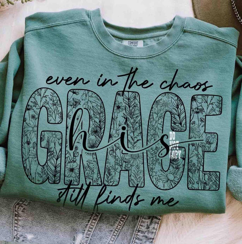 May include: A teal-colored sweatshirt with the words "even in the chaos GRACE his GRACE still finds me" in black script and floral design. The sweatshirt is made of a soft material and has a crew neck.