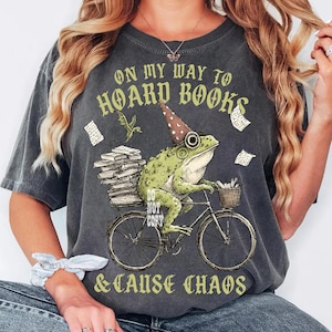 May include: Dark grey t-shirt featuring a frog on a bicycle, wearing a party hat and carrying books. The text reads "ON MY WAY TO HOARD BOOKS & CAUSE CHAOS". A small dragon and floating papers are also in the design.
