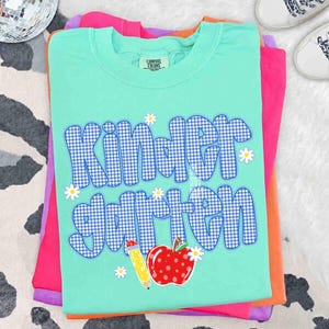以下が含まれることがあります： 青と白のギンガムチェック柄でKindergartenという文字がプリントされたミントグリーンの半袖Tシャツ。デザインには、黄色い鉛筆、白い水玉模様の赤いリンゴ、そして文字の周りに散りばめられた小さな白いデイジーの花が含まれています。