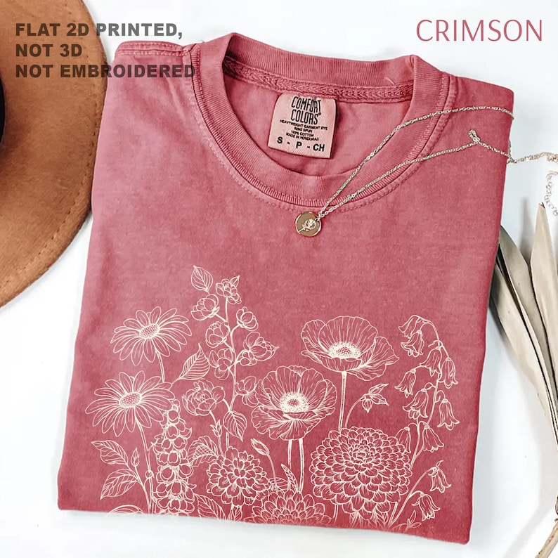 May include: Crimson-colored t-shirt with a white floral design. The shirt has a crew neck and short sleeves. The text "FLAT 2D PRINTED, NOT 3D, NOT EMBROIDERED" is printed on the top left. A silver necklace with a pendant is on the shirt.