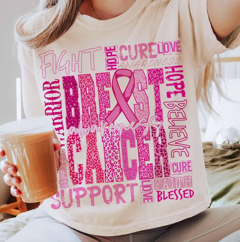 Op de afbeelding: Cr&egrave;mekleurig T-shirt met een roze borstkankerbewustzijnsontwerp. Het ontwerp bevat een roze lint, de woorden "Breast Cancer Awareness" en andere ondersteunende woorden in verschillende roze lettertypen. Het shirt is een casual stijl.