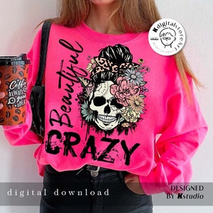 Peut inclure: Sweat-shirt rose avec un motif de crâne noir orné de fleurs et d'un chignon désordonné. Le texte "Beautiful Crazy" est imprimé sur le sweat-shirt.