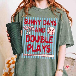 May include: Sage green t-shirt with the text "SUNNY DAYS AND DOUBLE PLAYS" in red. The design includes a baseball, a baseball bat, and a bow. The shirt is a comfortable, casual style.