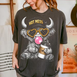 K&ouml;nnte beinhalten: Dunkelgraues T-Shirt mit einem Cartoon-Highland-Rind mit herzf&ouml;rmiger Sonnenbrille, einer Kaugummiblase und einer Kaffeetasse. Das Rind tr&auml;gt einen Hut mit dem Text "HOT MESS". Das Shirt hat eine lockere Passform.