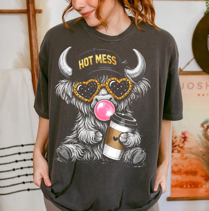 K&ouml;nnte beinhalten: Dunkelgraues T-Shirt mit einem Cartoon-Highland-Rind mit herzf&ouml;rmiger Sonnenbrille, einer Kaugummiblase und einer Kaffeetasse. Das Rind tr&auml;gt einen Hut mit dem Text "HOT MESS". Das Shirt hat eine lockere Passform.