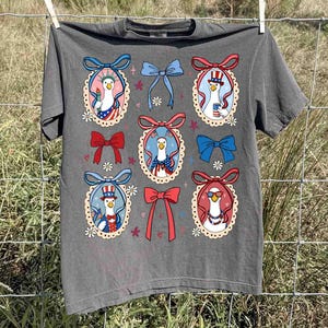 Op de afbeelding: Donkergrijs t-shirt met een herhaald patroon van cartoon ganzen in patriottische outfits, omlijst door decoratieve strikken en bloemaccenten. De ganzen worden afgebeeld met Amerikaanse vlagmotieven en accessoires, ter viering van Independence Day.