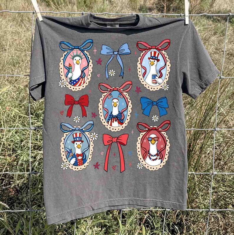 Op de afbeelding: Donkergrijs t-shirt met een herhaald patroon van cartoon ganzen in patriottische outfits, omlijst door decoratieve strikken en bloemaccenten. De ganzen worden afgebeeld met Amerikaanse vlagmotieven en accessoires, ter viering van Independence Day.