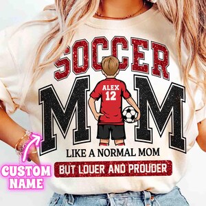 May include: Cream-colored t-shirt with the words "SOCCER MOM" in red glitter lettering above the word "MOM" in black block letters. A cartoon of a soccer player in a red jersey is in the center. The text "LIKE A NORMAL MOM BUT LOUDER AND PROUDER" is below.