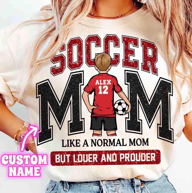 May include: Cream-colored t-shirt with the words "SOCCER MOM" in red glitter lettering above the word "MOM" in black block letters. A cartoon of a soccer player in a red jersey is in the center. The text "LIKE A NORMAL MOM BUT LOUDER AND PROUDER" is below.