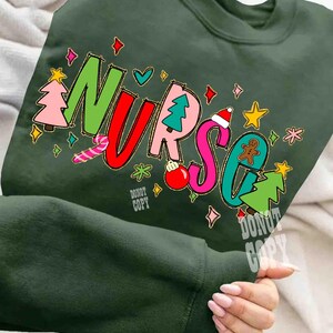 May include: Dark green sweatshirt with the word "NURSE" in colorful, festive lettering. The letters are decorated with Christmas-themed graphics, including a Christmas tree, gingerbread man, and candy cane. The words "Do Not Copy" are also visible.