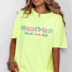 May include: A neon yellow t-shirt with a graphic that reads "beach book club" in blue script. The graphic includes illustrations of a flower, an open book, a crab, a stack of books, and a starfish, all in blue and pink.