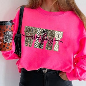 May include: A vibrant pink sweatshirt featuring the word "wifey" in a decorative font. The letters are designed with leopard print, black and white checkered patterns, and a tan and white checkered pattern. A black coffee cup is held in the left hand.