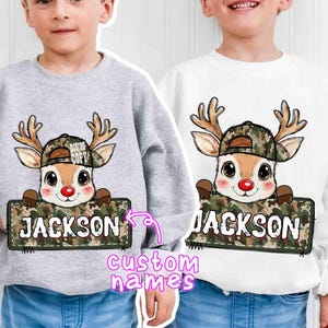 May include: Two sweatshirts, one gray and one white, each featuring a cartoon deer wearing a camouflage hat and holding a sign that says "JACKSON". The deer has a red nose and brown antlers. The sweatshirts are custom name designs.