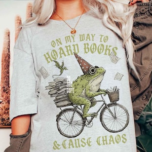 May include: Pale grey t-shirt featuring a frog on a bicycle, wearing a wizard's hat, and carrying books. The text reads "ON MY WAY TO HOARD BOOKS & CAUSE CHAOS" in green.