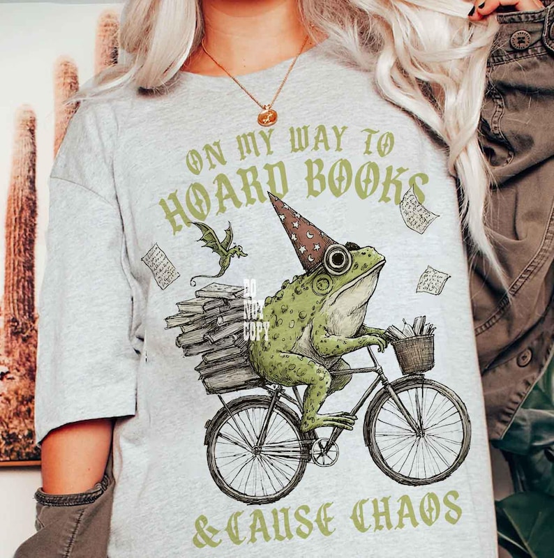 May include: Pale grey t-shirt featuring a frog on a bicycle, wearing a wizard's hat, and carrying books. The text reads "ON MY WAY TO HOARD BOOKS & CAUSE CHAOS" in green.
