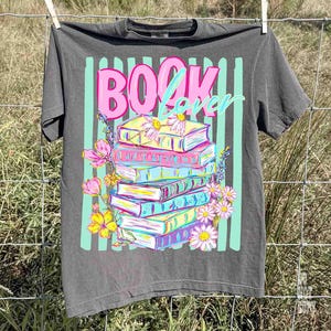 May include: A gray t-shirt with the words "BOOK Lover" in pink and teal. The graphic features a stack of colorful books with flowers. The shirt is hanging on a clothesline.