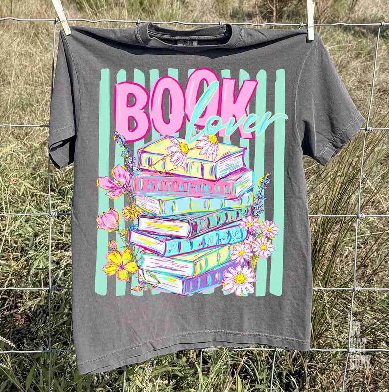May include: A gray t-shirt with the words "BOOK Lover" in pink and teal. The graphic features a stack of colorful books with flowers. The shirt is hanging on a clothesline.