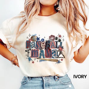 K&ouml;nnte beinhalten: Elfenbeinfarbenes T-Shirt mit einem Baseball-Motiv. Das Design zeigt die Worte "Baseball Mama" in Rot, Wei&szlig; und Blau, zusammen mit Baseball-bezogenen Illustrationen wie einem Schl&auml;ger, Sternen und einem Baseball.