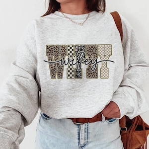 May include: A light grey sweatshirt featuring the word "wifey" in a decorative font. The letters are filled with leopard print, checkered, and polka dot patterns. A brown leather belt and a brown shoulder bag are also visible.