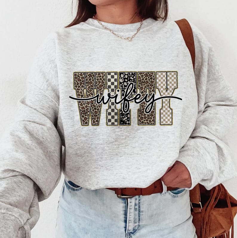 May include: A light grey sweatshirt featuring the word "wifey" in a decorative font. The letters are filled with leopard print, checkered, and polka dot patterns. A brown leather belt and a brown shoulder bag are also visible.