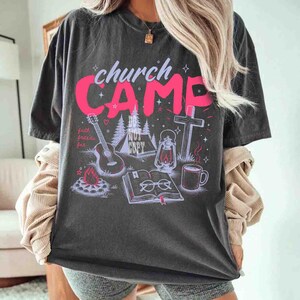 K&ouml;nnte beinhalten: Dunkelgraues T-Shirt mit einem rosa Grafikdesign mit der Aufschrift "church CAMP", zusammen mit Illustrationen einer Gitarre, eines Zeltes, eines Kreuzes, einer Laterne, einer Kaffeetasse und eines offenen Buches. Das Shirt hat auch den Text "faith, friends, fun".