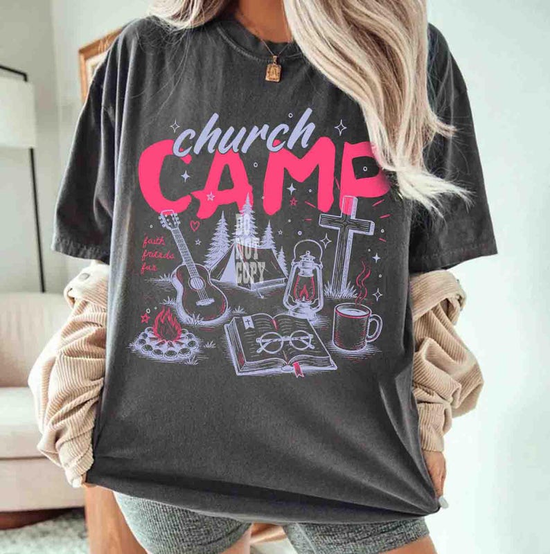 K&ouml;nnte beinhalten: Dunkelgraues T-Shirt mit einem rosa Grafikdesign mit der Aufschrift "church CAMP", zusammen mit Illustrationen einer Gitarre, eines Zeltes, eines Kreuzes, einer Laterne, einer Kaffeetasse und eines offenen Buches. Das Shirt hat auch den Text "faith, friends, fun".