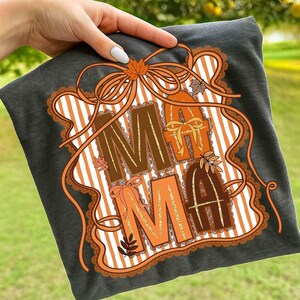 Op de afbeelding: Donkergrijs t-shirt met het woord "MAMA" in bruine en oranje letters. Het ontwerp bevat herfstthema-elementen zoals bladeren en strikken, tegen een gestreepte achtergrond. Het ontwerp is omlijst met een decoratieve rand.