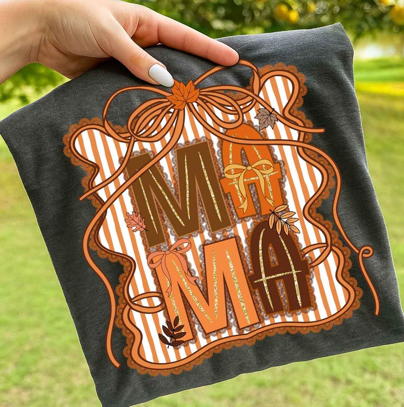 Op de afbeelding: Donkergrijs t-shirt met het woord "MAMA" in bruine en oranje letters. Het ontwerp bevat herfstthema-elementen zoals bladeren en strikken, tegen een gestreepte achtergrond. Het ontwerp is omlijst met een decoratieve rand.