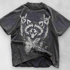 Pu&ograve; includere: T-shirt grigio scuro con una grafica bianca invecchiata di un procione che fa il gesto del rock and roll. Il procione ha la lingua di fuori. La maglietta ha un aspetto vintage e usurato.