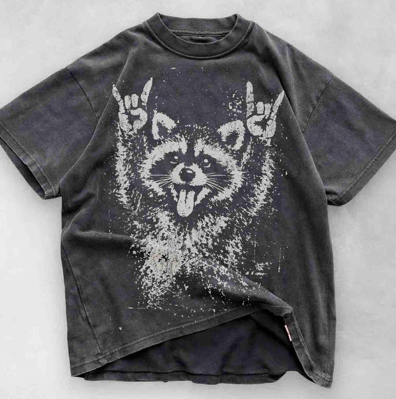 Pu&ograve; includere: T-shirt grigio scuro con una grafica bianca invecchiata di un procione che fa il gesto del rock and roll. Il procione ha la lingua di fuori. La maglietta ha un aspetto vintage e usurato.