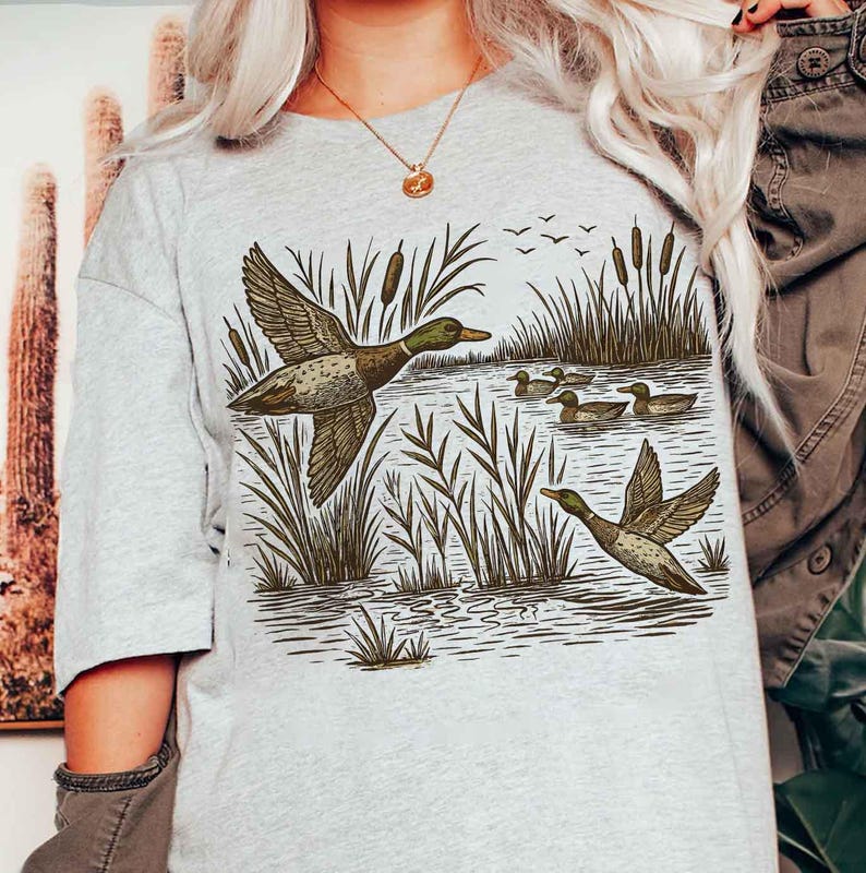 May include: A light gray t-shirt featuring a detailed illustration of ducks in a wetland environment. The design includes ducks in flight and swimming, with cattails and water plants. The artwork is in shades of brown and green.