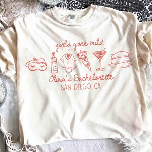 May include: Cream-colored t-shirt with red graphic text and illustrations. The text reads "girls gone mild" above illustrations of a sleep mask, wine bottle, pajamas, flowers, a cocktail, and pillows. Below, it says "Olivia's Bachelorette SAN DIEGO, CA."