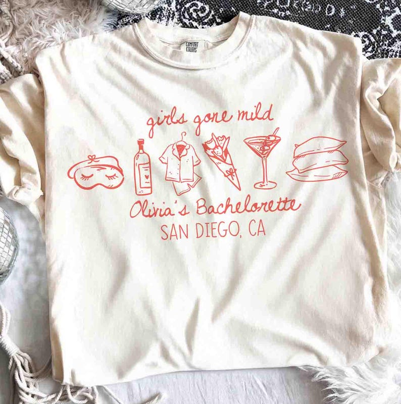 May include: Cream-colored t-shirt with red graphic text and illustrations. The text reads "girls gone mild" above illustrations of a sleep mask, wine bottle, pajamas, flowers, a cocktail, and pillows. Below, it says "Olivia's Bachelorette SAN DIEGO, CA."