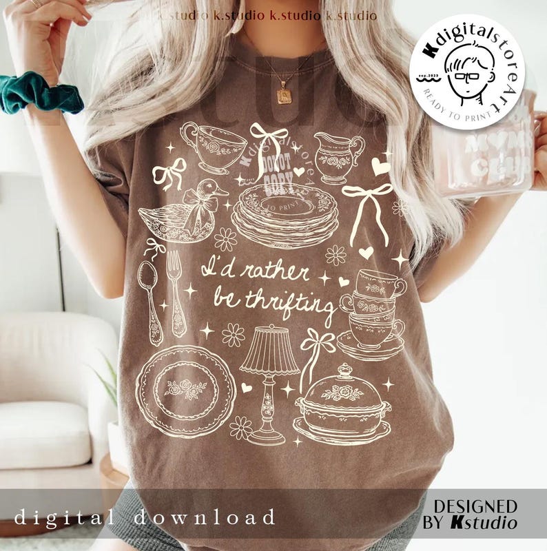 May include: Brown t-shirt with a white graphic design featuring vintage kitchenware illustrations and the text "I'd rather be thrifting." The design includes teacups, plates, a duck, and a lamp. "Digital Download" and "Designed by Kstudio" are also printed on the shirt.