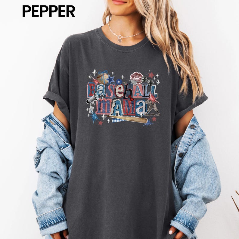 K&ouml;nnte beinhalten: Dunkelgraues T-Shirt mit dem Text "Baseball Mama" in einem farbenfrohen, verwaschenen Design. Die Grafik enth&auml;lt Sterne, einen Baseballschl&auml;ger und einen Baseball. Das Shirt hat kurze &Auml;rmel und eine lockere Passform.