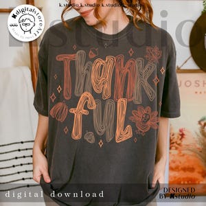 K&ouml;nnte beinhalten: Ein anthrazitfarbenes Kurzarm-T-Shirt mit dem Schriftzug Thankful in einer stilisierten orange- und braunfarbenen Linien-Schriftart. Das Design enth&auml;lt grafische Illustrationen eines Truthahns, eines K&uuml;rbisses, von Eicheln und Ahornbl&auml;ttern.