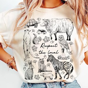 May include: Cream-colored sweatshirt featuring a black and white illustration of various animals, including a zebra, owl, and rabbit. The text "Respect the local" is written in a script font. The design includes leaves and acorns.