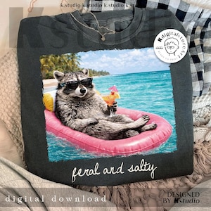 May include: Dark gray t-shirt featuring a raccoon wearing sunglasses and floating on a pink inflatable in the ocean, holding a cocktail. The text "feral and salty" is below the image. The words "digital download" and "designed by kstudio" are also visible.