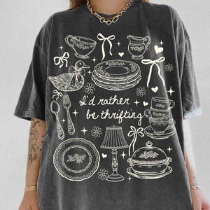 May include: Dark gray t-shirt with a whimsical design featuring teacups, plates, a duck, and the text "I'd rather be thrifting." The design is in white, creating a vintage aesthetic. The shirt has short sleeves and a relaxed fit.