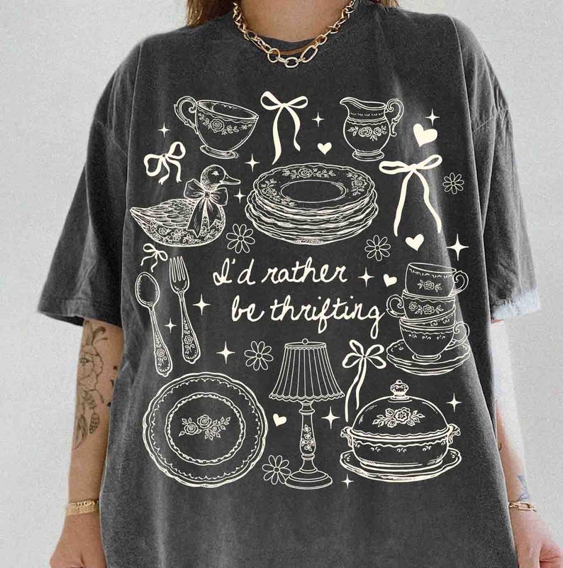 May include: Dark gray t-shirt with a whimsical design featuring teacups, plates, a duck, and the text "I'd rather be thrifting." The design is in white, creating a vintage aesthetic. The shirt has short sleeves and a relaxed fit.