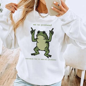 May include: White sweatshirt featuring a cartoon frog with the text "me: no problemo!" and "narrator: but it was all problemo". The frog is green with a light green belly and is raising its arms.