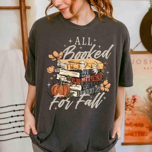 May include: Dark gray t-shirt with a fall-themed graphic. The design features a stack of books, a pumpkin, and floral accents, with the text "ALL Booked For Fall" in a decorative font. The shirt has a crew neck and short sleeves.