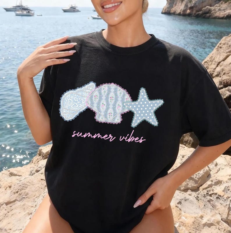 May include: Black t-shirt with a seashell, floral, and starfish design in light blue and pink. The words "summer vibes" are written in pink script below the design. The shirt is worn outdoors near the ocean.