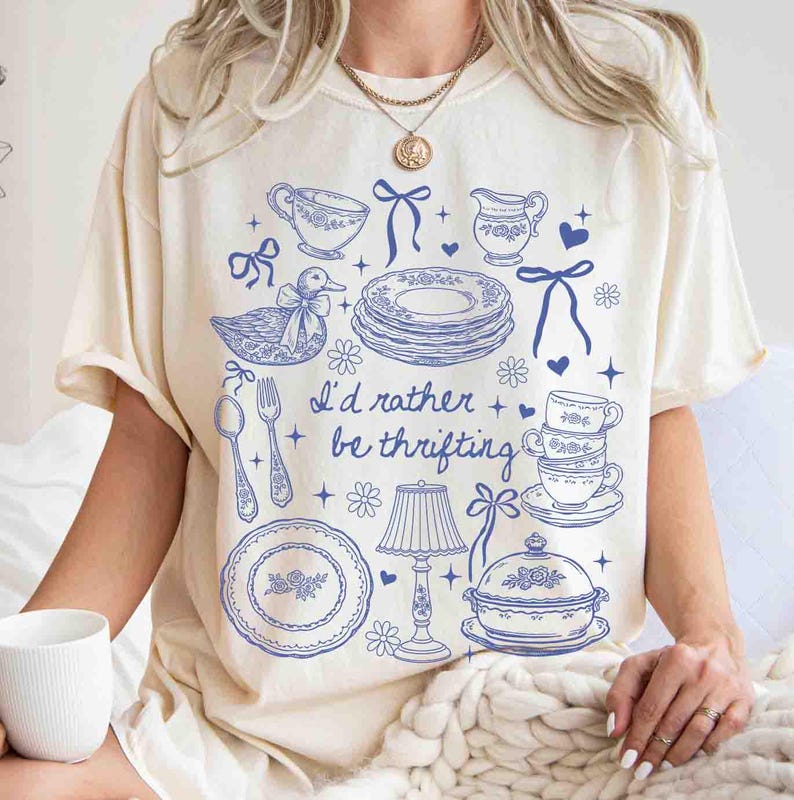 May include: Cream-colored t-shirt featuring a blue illustration of vintage tableware, including plates, cups, and a duck. The text "I'd rather be thrifting" is printed below the illustrations. The shirt has short sleeves.