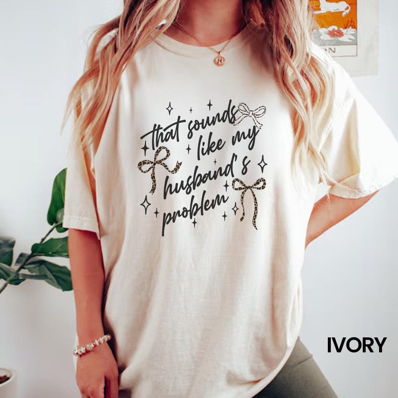 May include: Ivory-colored t-shirt with the text "That sounds like my husband's problem" in a black script font. The text is decorated with leopard print bows and star accents. The shirt is a relaxed fit.