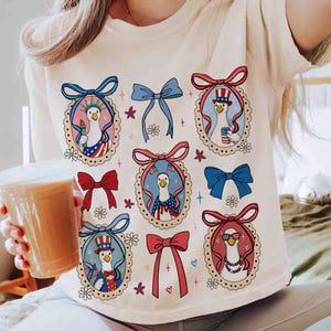 Op de afbeelding: Cr&egrave;mekleurig T-shirt met een grillig ontwerp van patriottische ganzen. Elke gans is omlijst in een decoratief ovaal, versierd met strikken en bloemmotieven. De ganzen zijn gekleed in rood, wit en blauw, met Amerikaanse vlaggen en vrijheidsfakkels.